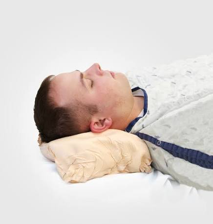 A man lying down with his eyes closed, resting his head on a beige crinkled pillow, wrapped in a white blanket with a navy blue collar.
